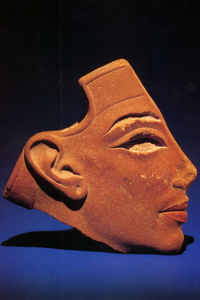 A studio photograph of an ancient Egyptian red terracotta or quartzite face profile fragment, set against a dark blue background with dramatic side lighting.